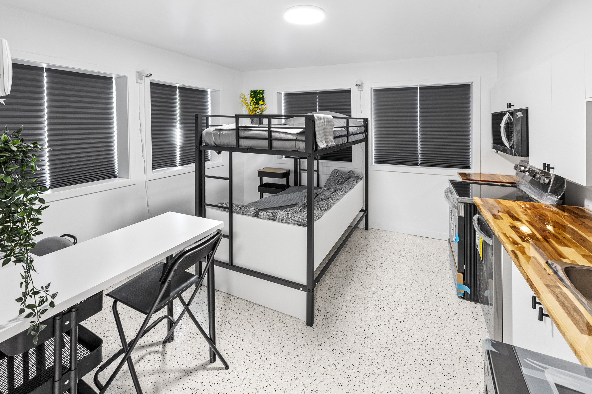 Unit 09 loft zone: welded steel loft frame with an adult twin hybrid gel mattress above, a full daybed with hybrid gel mattress below, blackout cellular shades on two windows, a mobile bamboo-top cart, and the kitchen counter visible at the right edge.